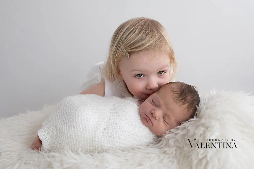 Toddler girl gently cuddling her newborn sibling on a fluffy white blanket- sibling portrait celebrating March babies