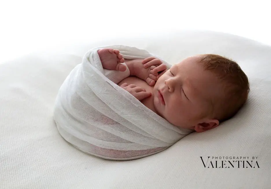 Newborn baby curled up on his back holding his little foot. He's laying on a white blanket and wrapped in a white blanket during a December newborn photoshoot in London.