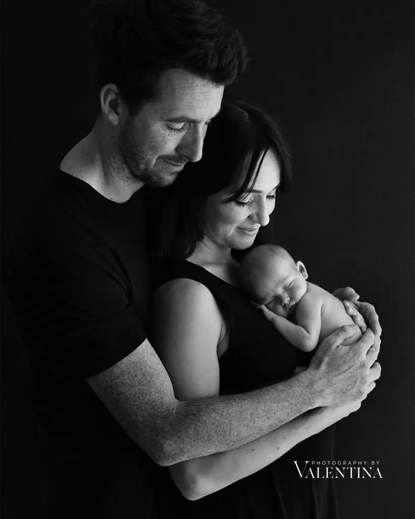Why Hiring a Professional Newborn Photographer Matters: black and white portrait of new parents lovingly holding their newborn baby in a calm and intimate studio setting by Photography by Valentina.