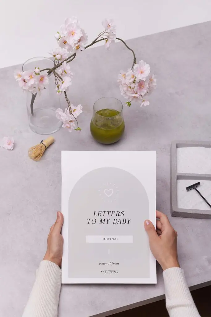 Hands holding the Letters to My Baby journal on a soft-toned table with cherry blossoms and a glass of matcha, evoking a calm spring mood—perfect for mums expecting a July baby.