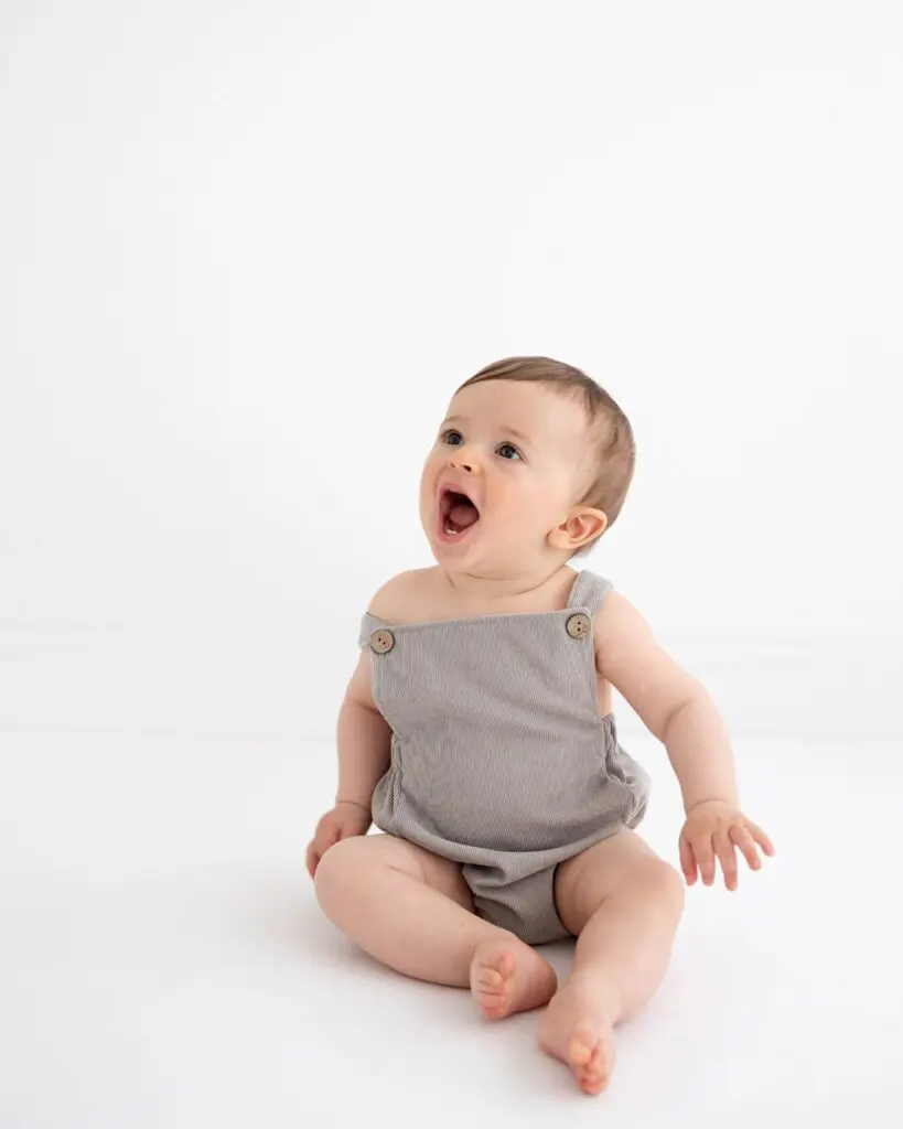 Laughing baby in a grey romper sitting against a white background – minimal studio portrait by Photography by Valentina