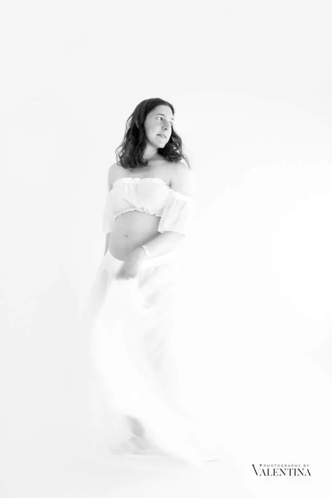 Elegant black and white maternity photography showing pregnant woman in flowing white dress during professional photo shoot for pregnant woman
