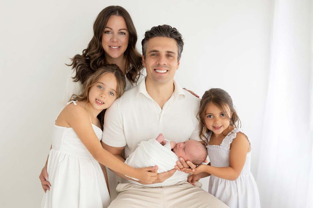 Family of five smiling during a newborn photoshoot in a bright London studio. Parents and two sisters wearing white outfits hold their newborn baby, creating a soft and timeless family portrait.