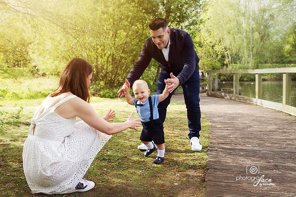 family portrait of mum, dad and baby in a park. Baby is walking towards mum and dad is helping him on his first steps