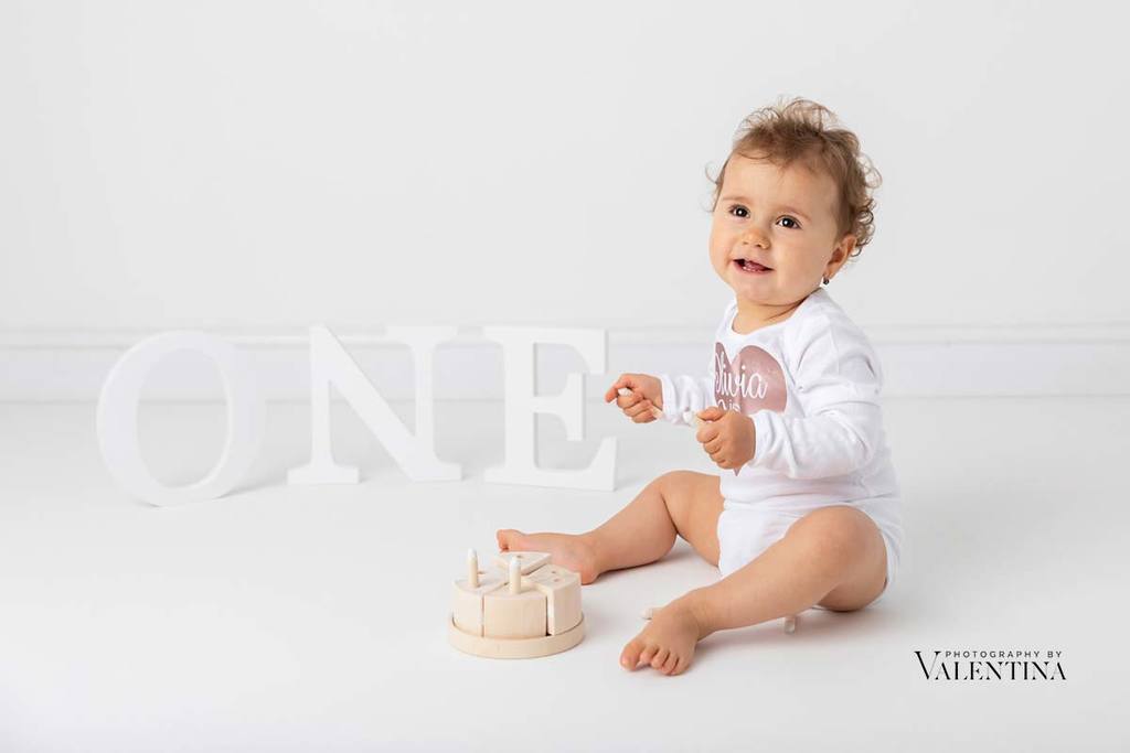Baby girl turning one! She sits on the floor with a wooden cake and the one words at the back