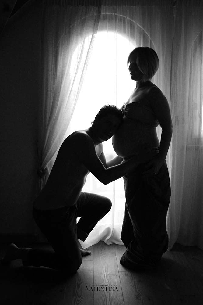silhouette of pregnant couple. Mum is standing in front of the window and dad is kneeling down with his ear on the pregnant belly. Black and white image.