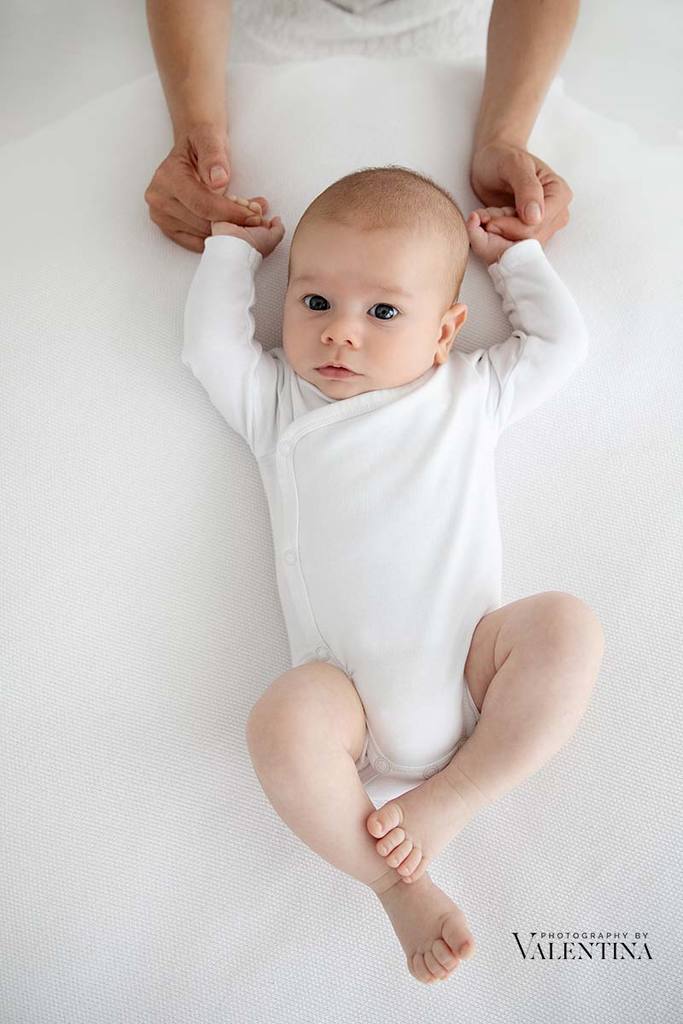 photo of 2 month old boy wearing a white bodysuit and laying on a beanbag. Mum is holding his hands.