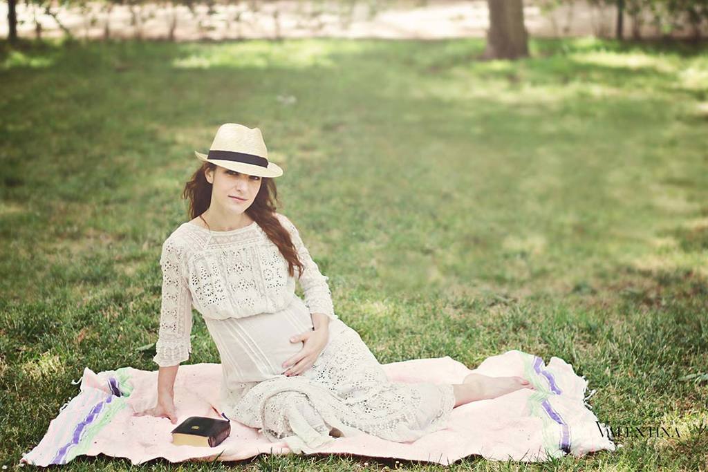 Pregnant woman sitting on the grass in a park. She is wearing a hat and holding her hand on the belly