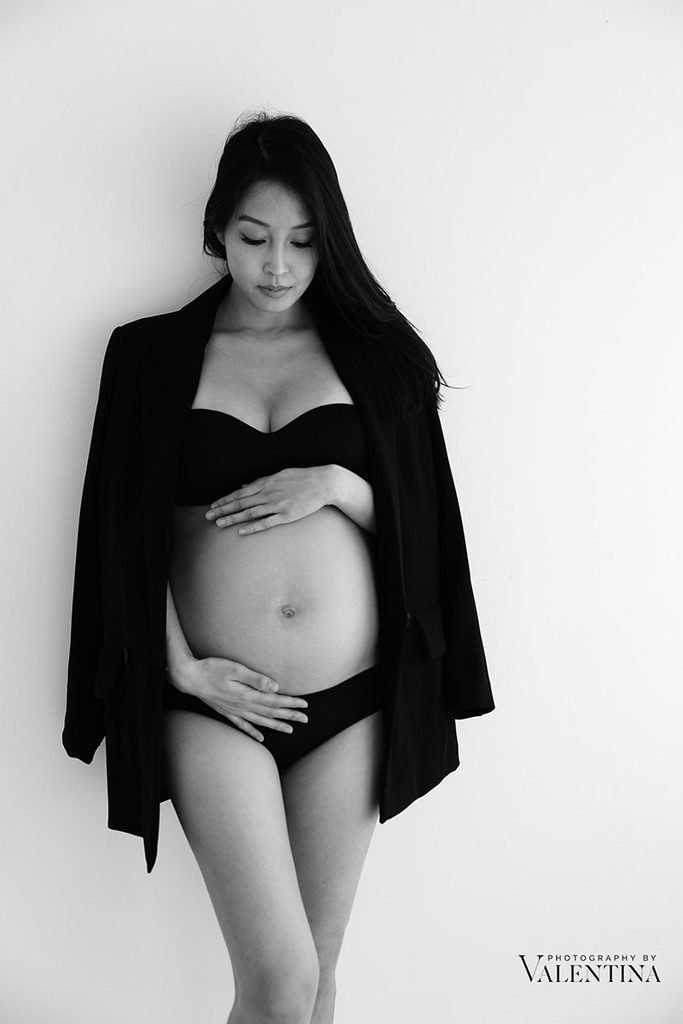 Pregnant mum is wearing her black lingerie and wearing a black blazer on her shoulder. She is leaning on the wall with her hands around the belly. Photo taken during a maternity photoshoot in London