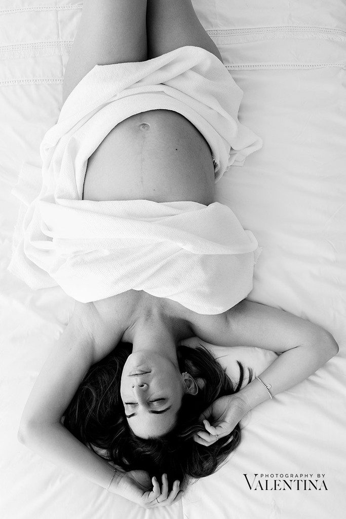 Pregnant mum laying on her back on a white bad. She is holding her arm up near her head. Photo taken by Valentina during a photoshoot.