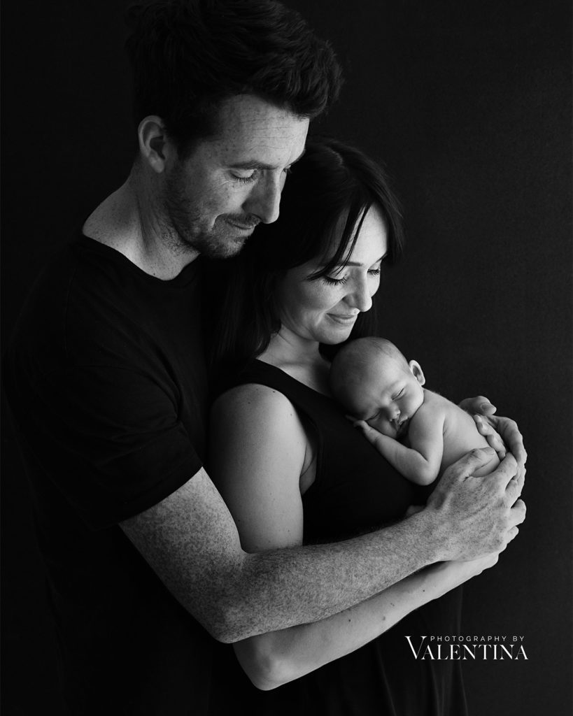 Stunning black and white family portrait. Mum is holding her newborn baby on her chest  while dad is behind them both.