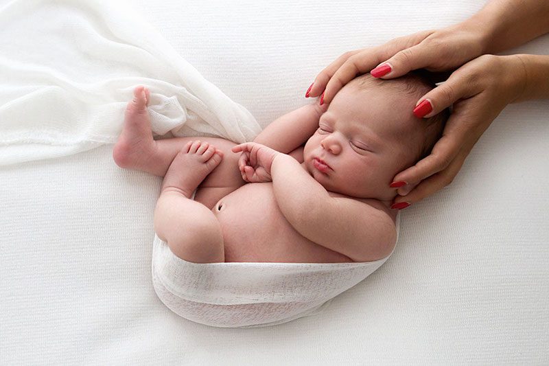 Newborn photographer in London – baby sleeping on white blanket, gently held by mum’s hands with red nails.