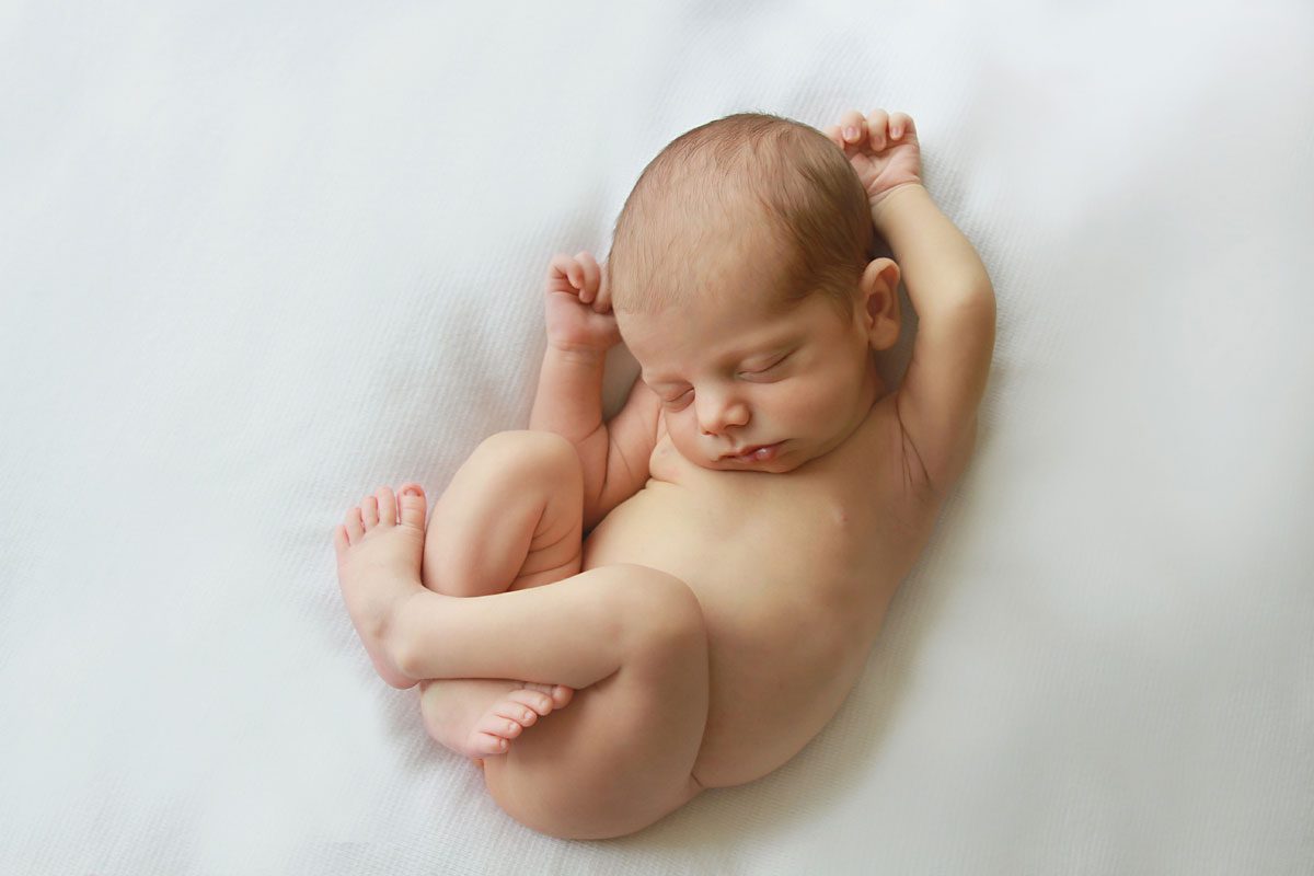newborn sleeping on a white blanket. He's naked and he's holding his arms up while is curling up his legs. Photo taken during a newborn photo shoot with London newborn photographer Valentina